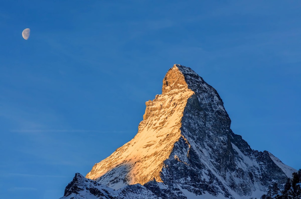 The Matterhorn glowing in warm light, with the Moon in the blue sky beside it